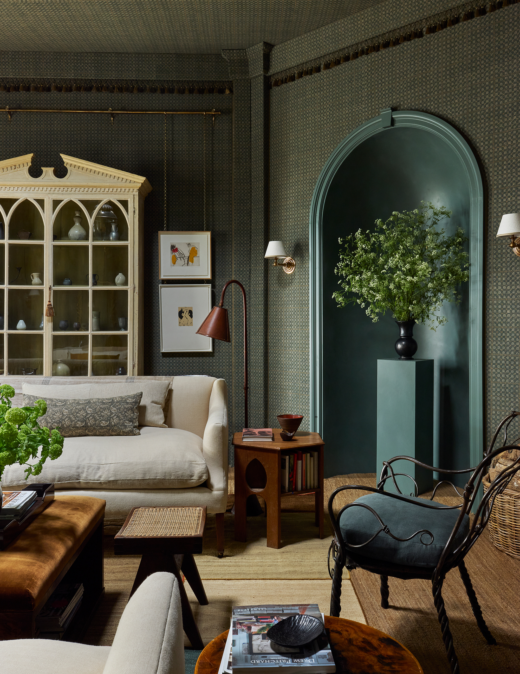 room with textured wallcovering, blue arch alcove with pedestal and vase of branches, white sofa, iron armchair, timber side table, stool, plush ottoman coffee table, jute carpet, and pictures on rail