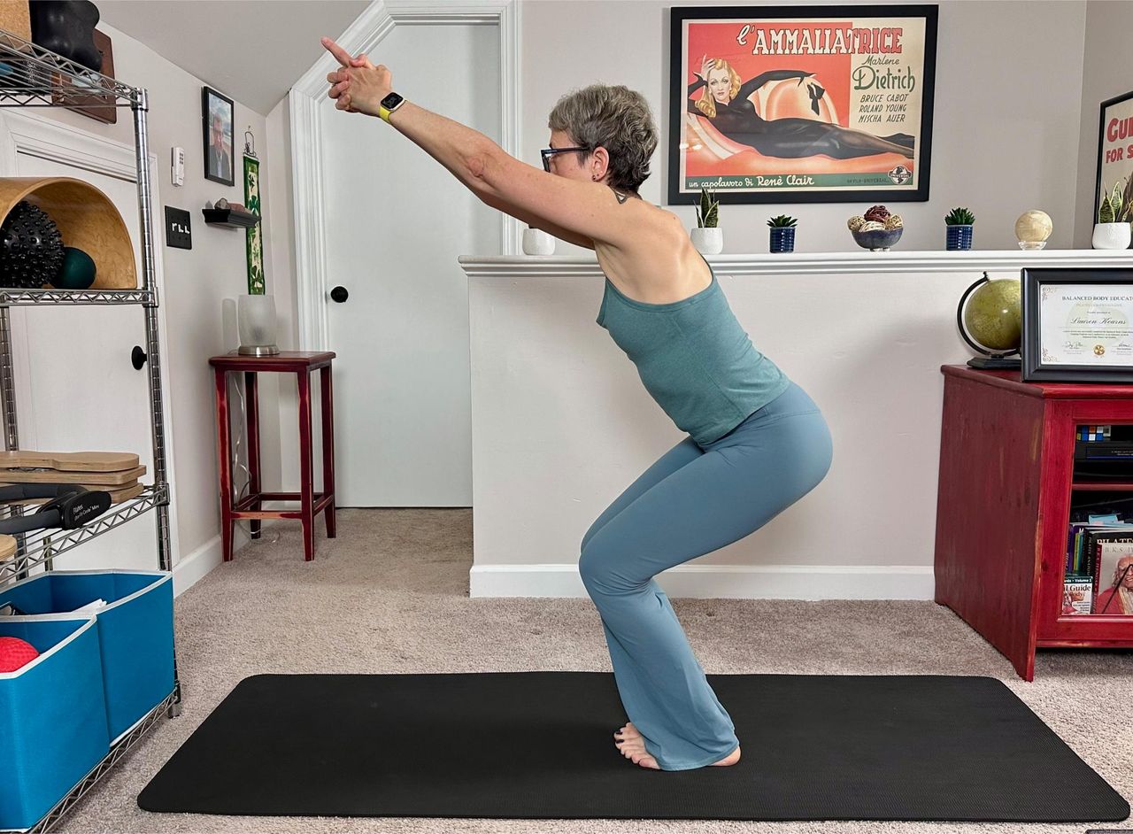 woman performing a chair pose in a living room setting on a black yoga mat wearing blue leggings and vest.