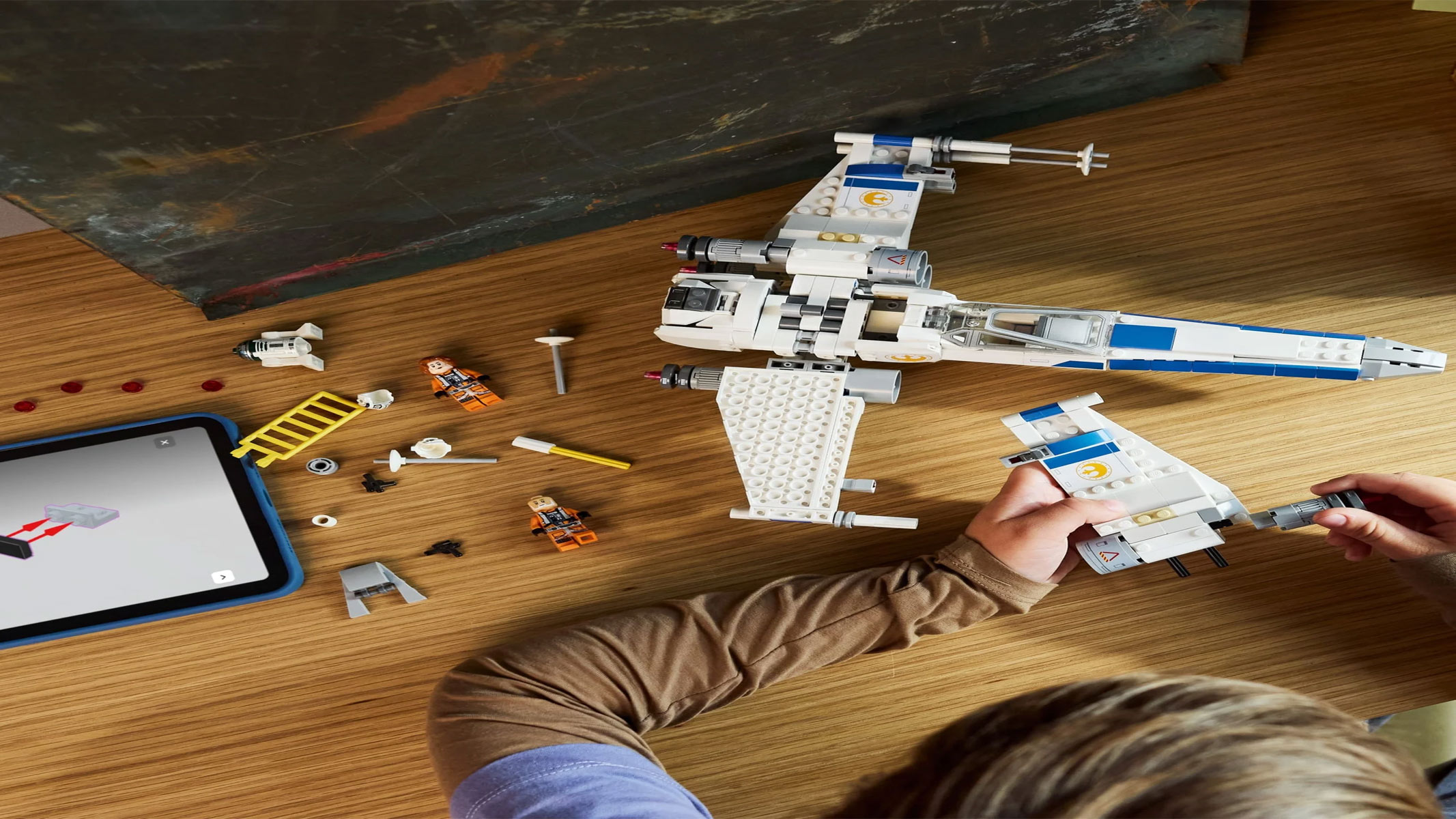 A child is in the process of building a brick-built spaceship, which is white and blue, on a brown wooden surface.