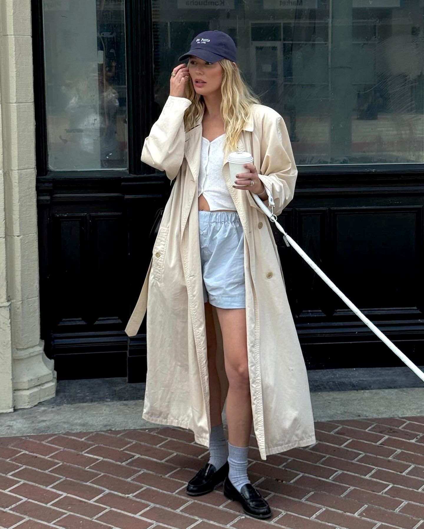 style influencer Megan Adelaide Vega poses on a brick sidewalk holding a cup of coffee wearing a blue baseball cap, long trench coat, white button-front top, blue boxer shorts, crew socks, and loafers