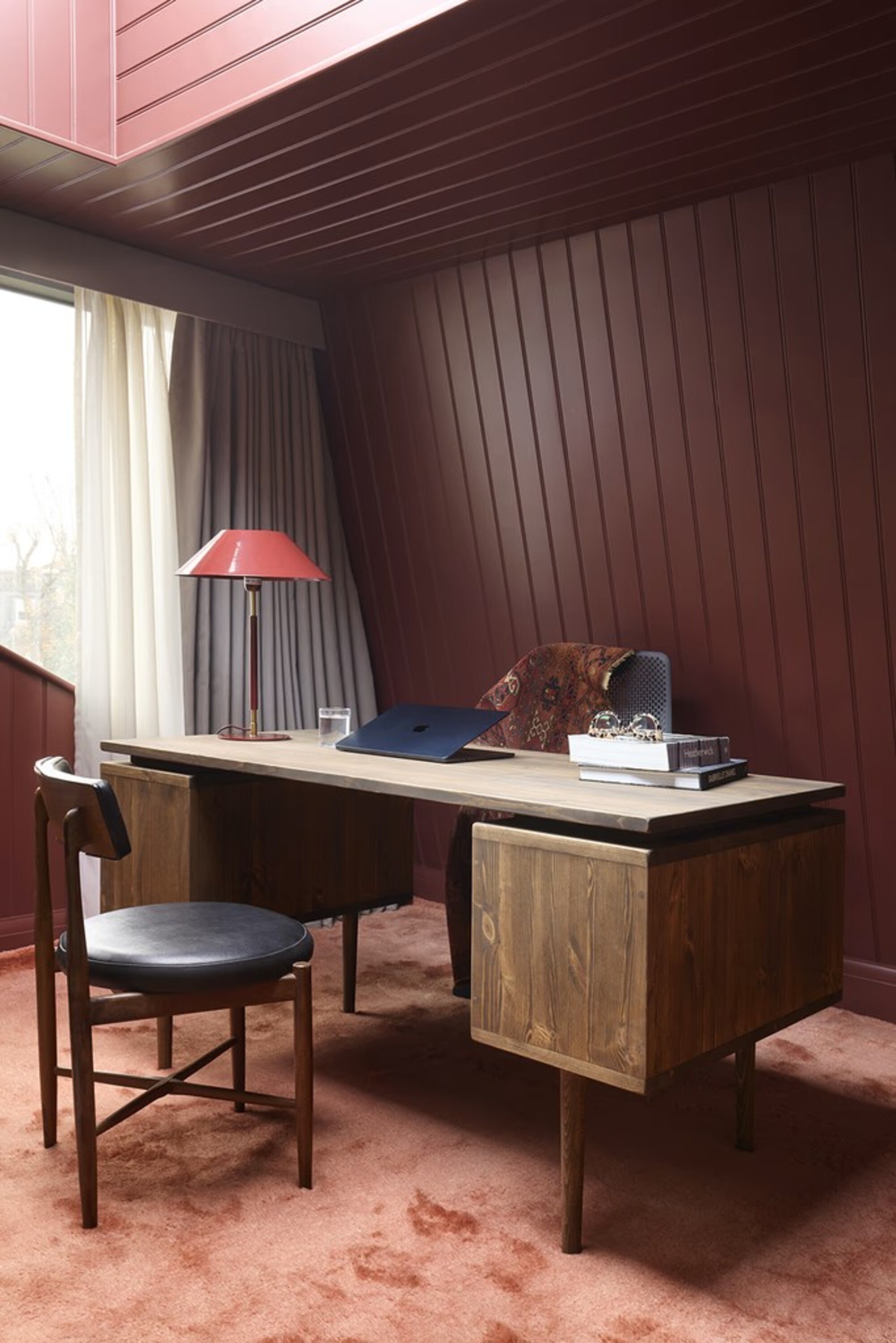 Image of a retro home office with pink carpets and berry-colored panel drenched walls and ceilings. there is a mid-century modern wooden desk in the center with a chair on either side.