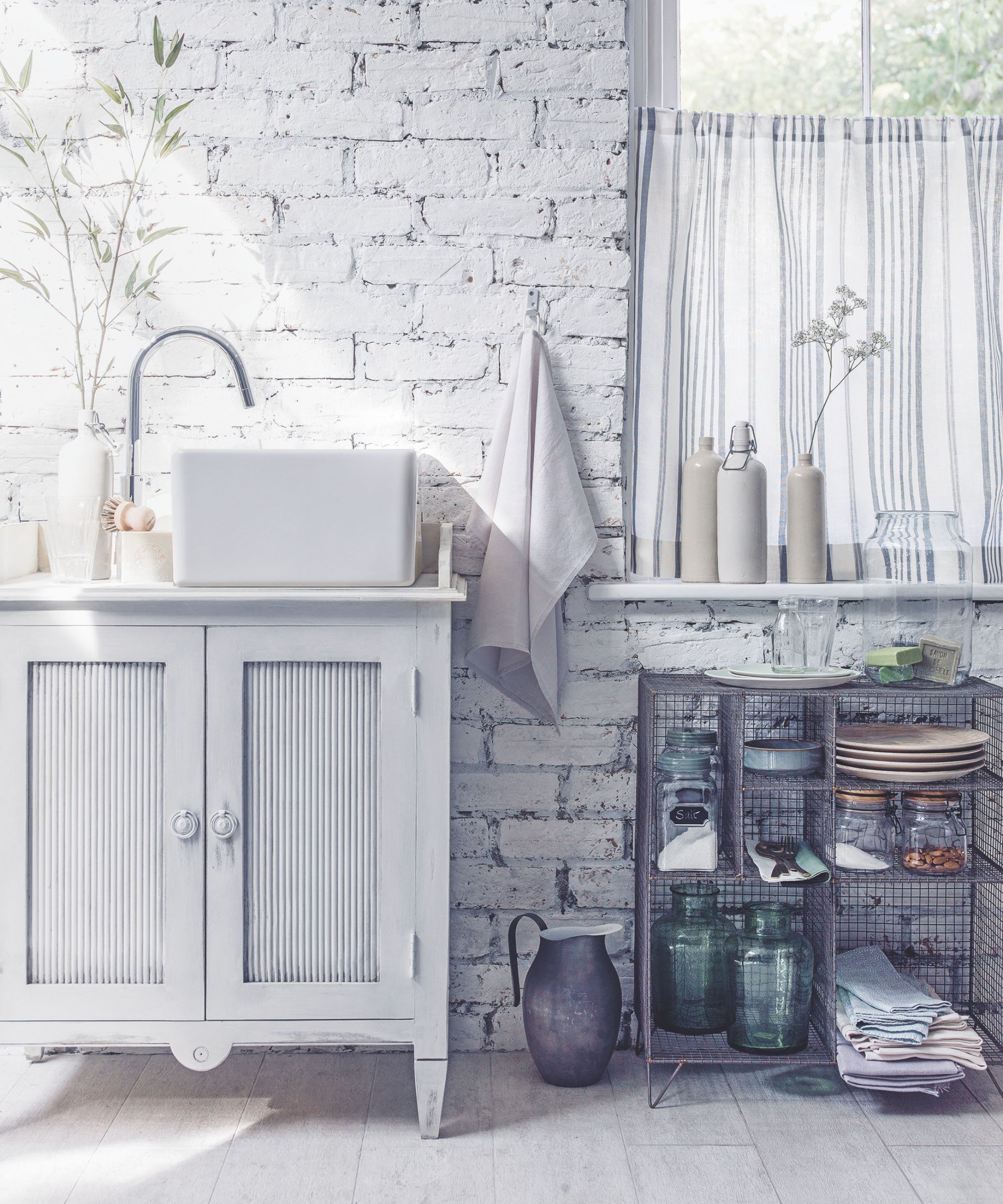 A whitewashed garden room with painted exposed brick walls, a large white cabinet, white sink, silver faucet and metal shelving.