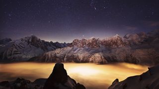 A stunning night landscape of snow-covered mountains under a starry sky, with glowing clouds illuminated by distant lights