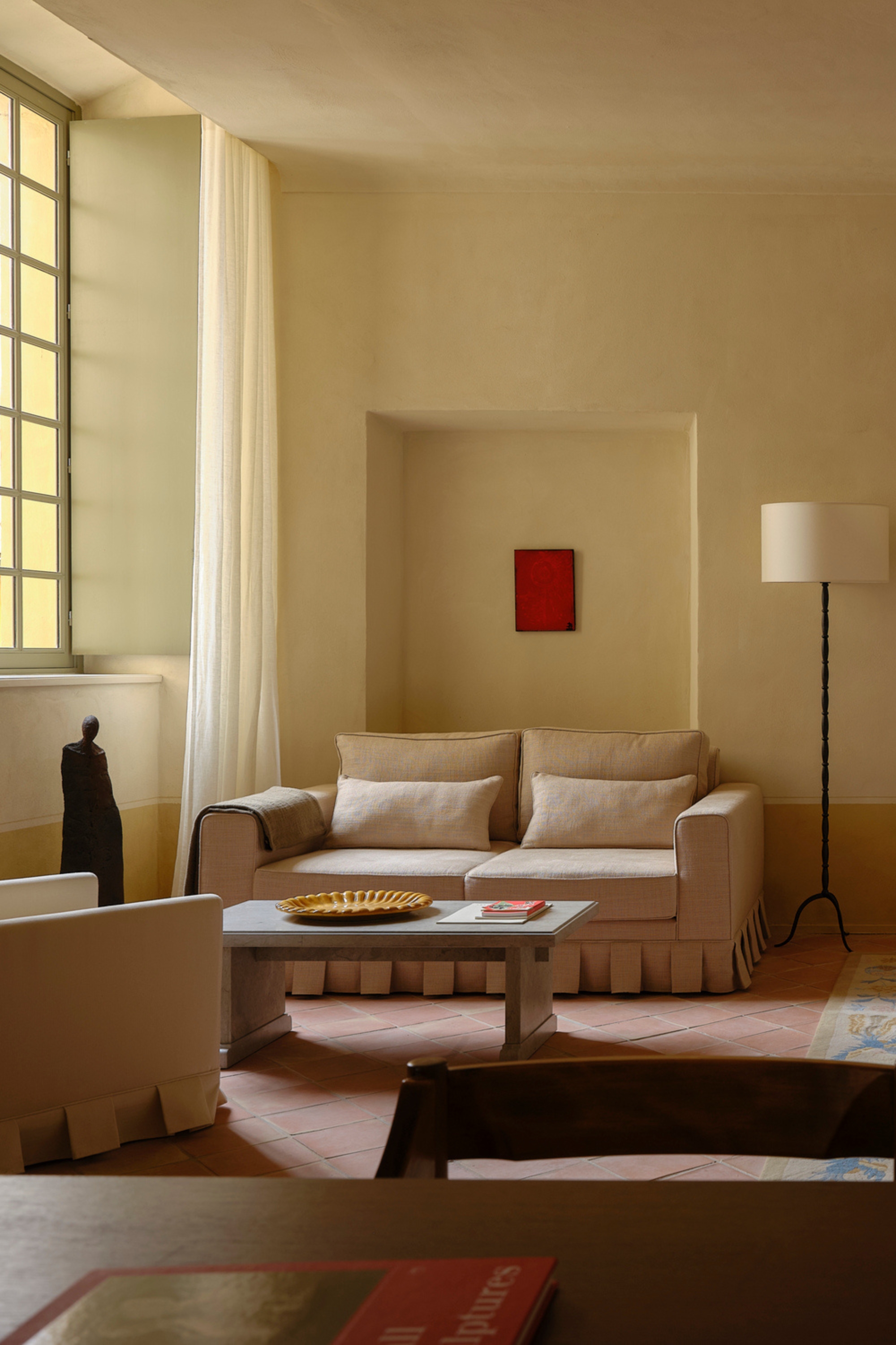 Beige, soft pink, and ochre living room with a skirted sofa, a stone coffee table, a large window with thin, flowy curtains hanging down, and a black iron floor lamp beside the sofa.