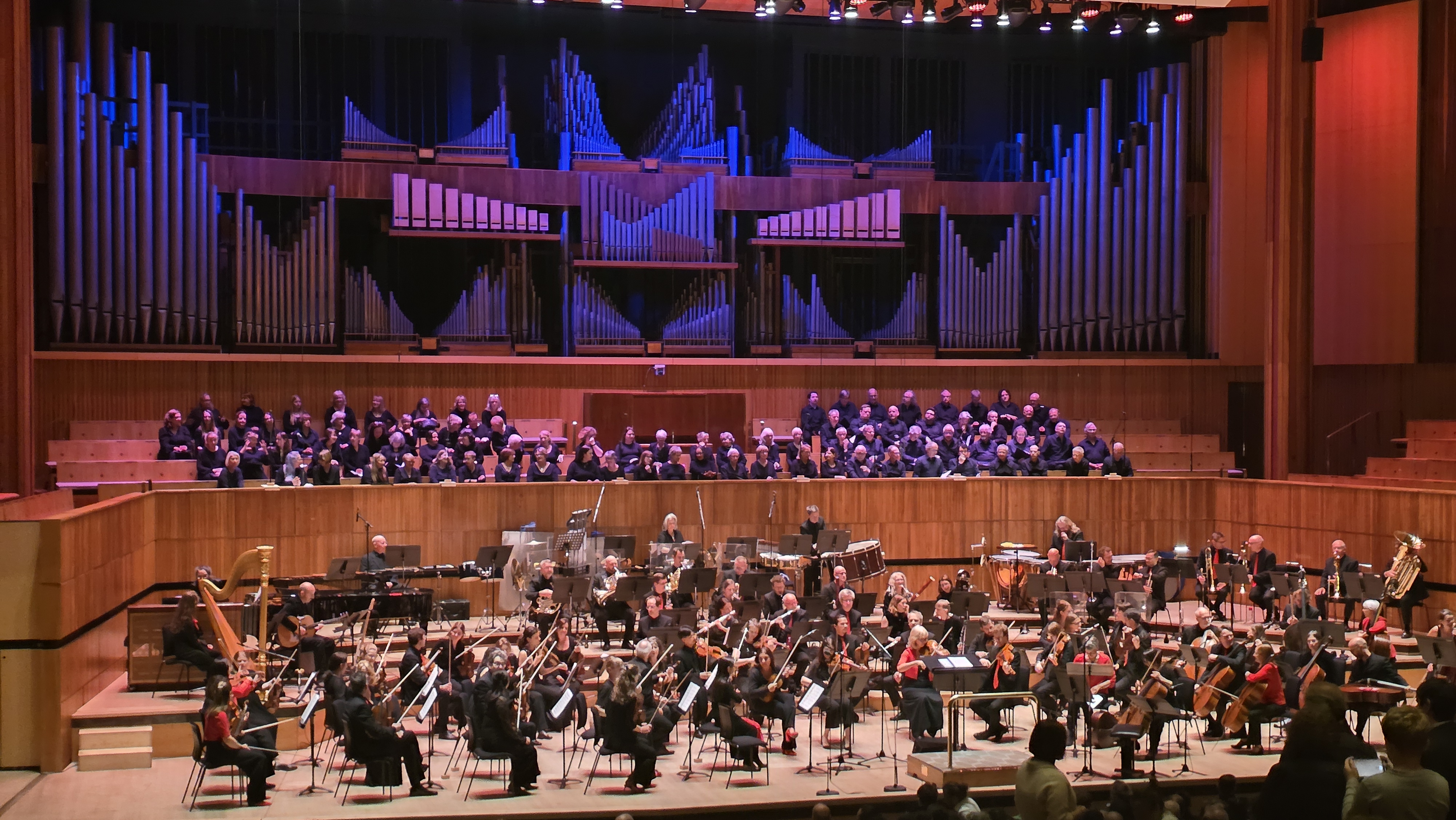 Stage full of musicians with string instruments, in the background there is a choir.