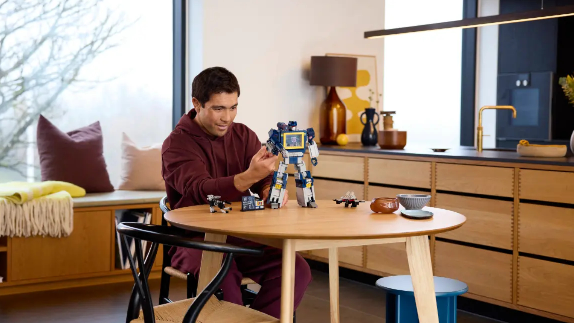 A smiling man sat at a table in a kitchen moves the arm of the Lego Transformers Soundwave model