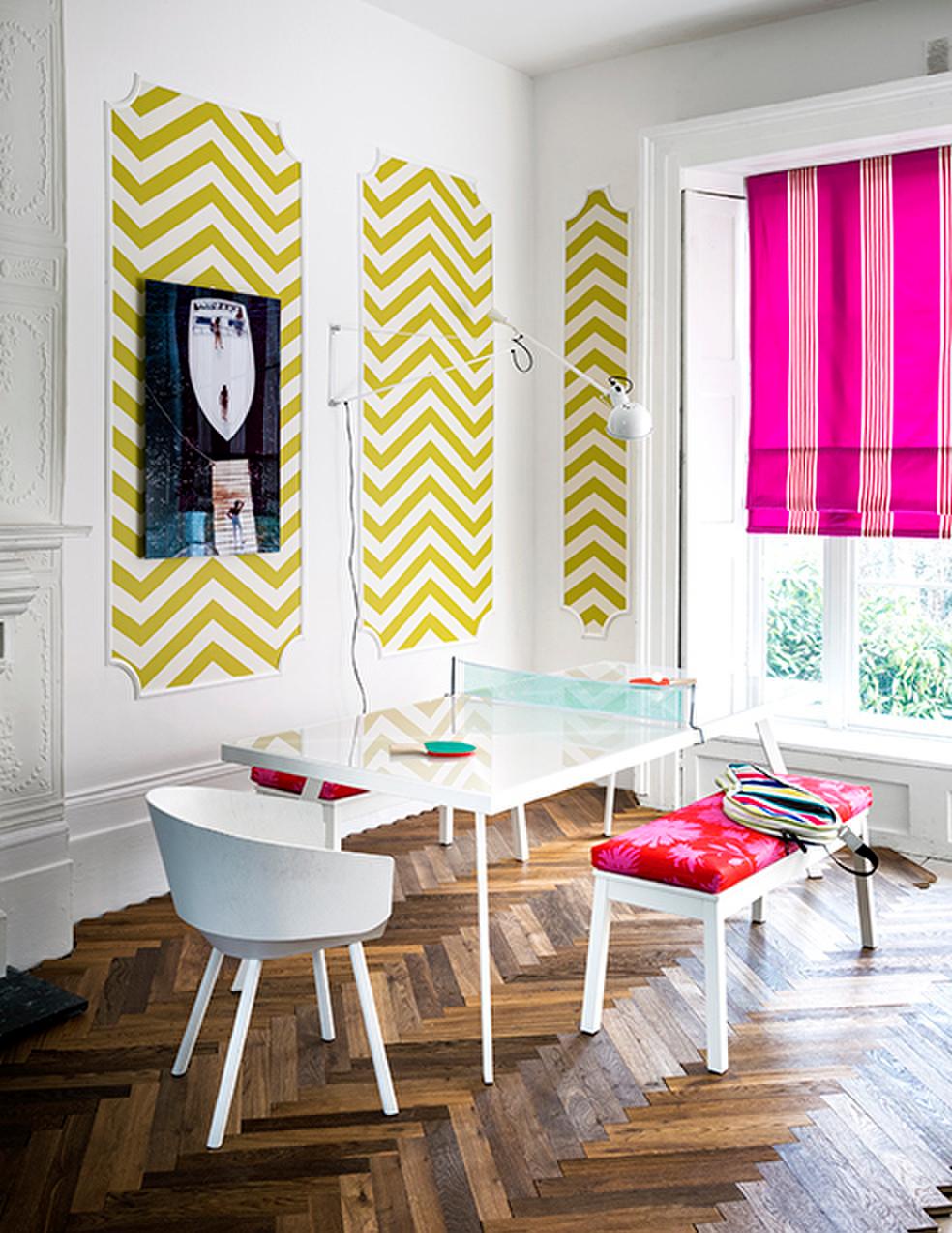 a white room with wooden flooring. There is a white wall with chevron wallpaper in panels on the wall. There is a white ping pong table with a hot pink bench seat.