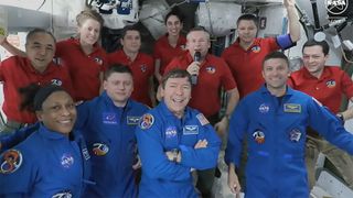 four astronauts in blue flight suits float in front of seven others in red shirts in a cramped, equipment-filled laboratory