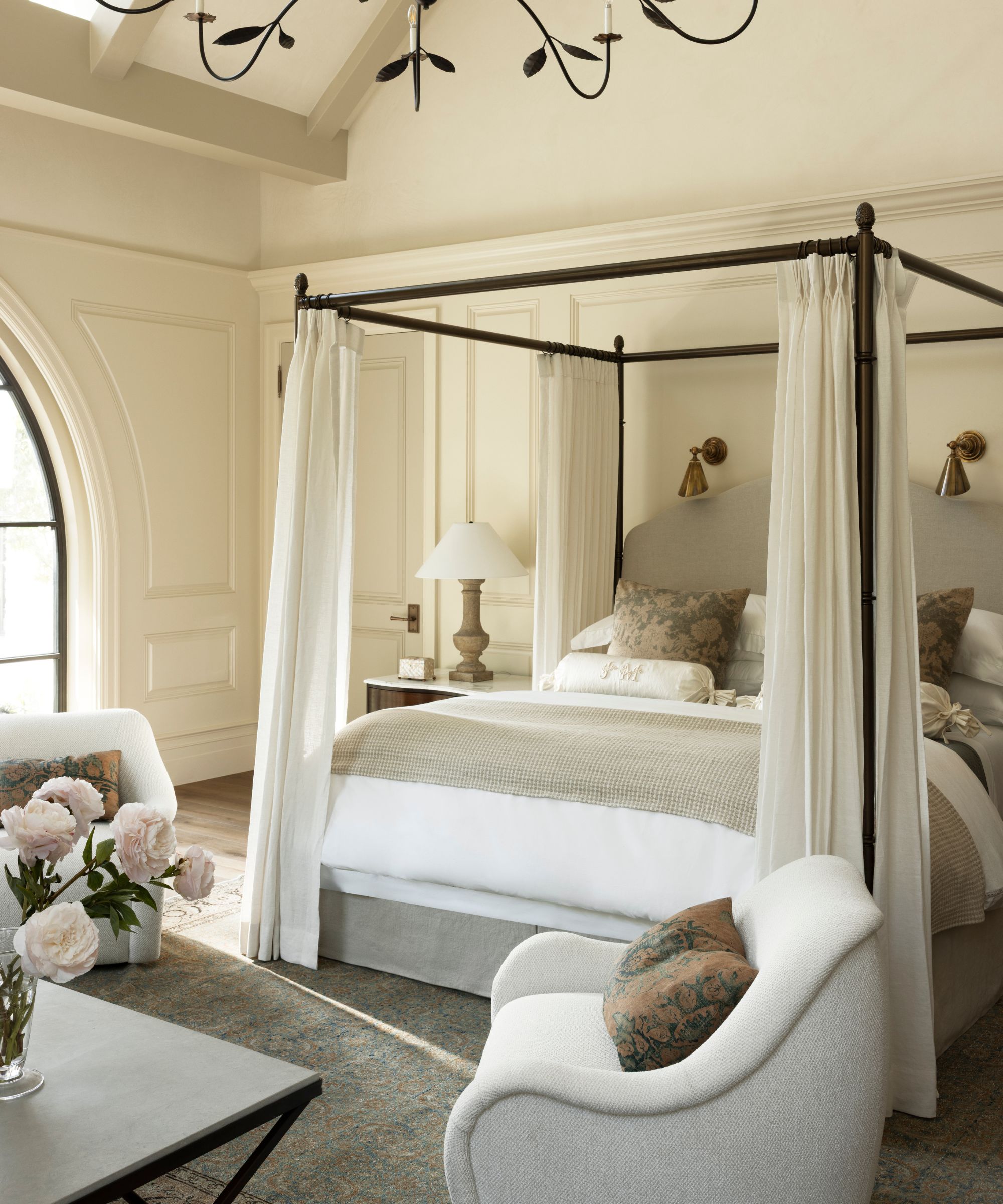 a neutral primary bedroom with a metal four poster bed with drapes and a small seating area
