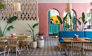 Pink arched walls with a view of the reception in the background. A mix of Blue sofas and wooden chairs with wooden rectangle tables. Cream hanging lights and green plants in pots.