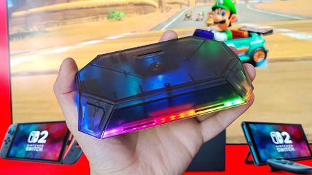 Hand holding Jsaux RGB docking station with Switch 2 console in backdrop and Mario Kart World footage on TV featuring Luigi.