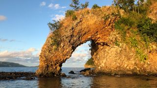 We see a natural arch rock along the shore by the water.