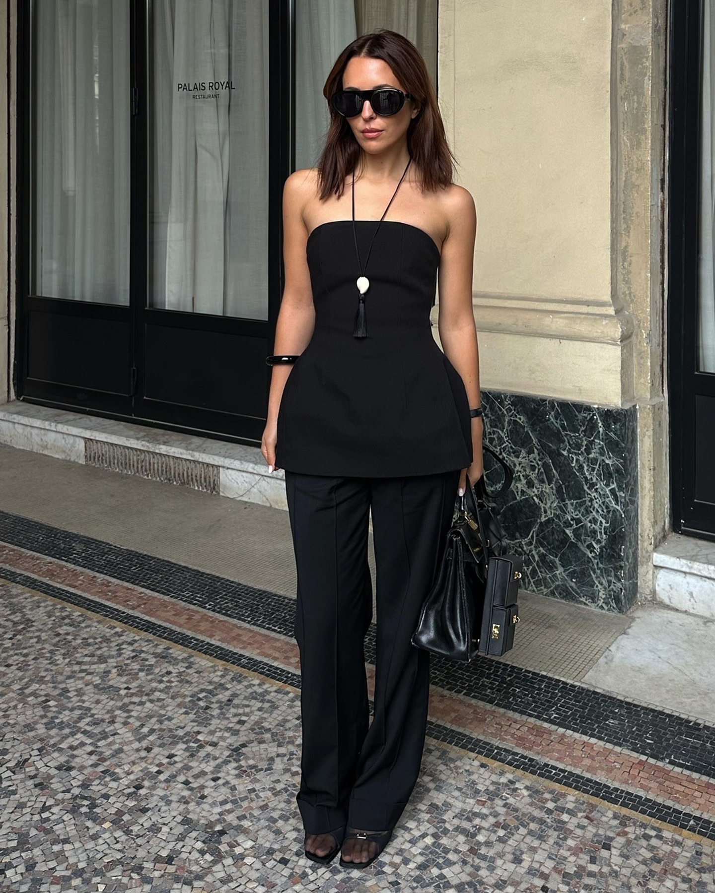 Style influencer Debora Rosa poses in a chic all-black summer 2026 outfit featuring sunglasses, a long cord pendant necklace, a sleeveless peplum top, a bag, pants, and mesh shoes.