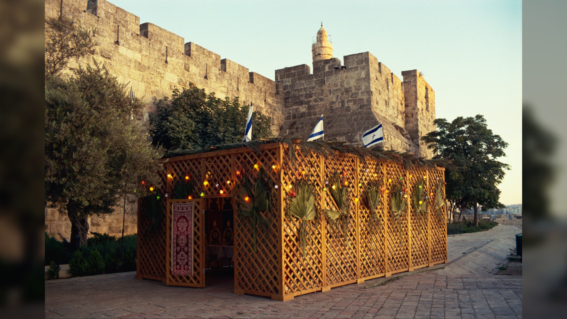 Here we see a Sukkot (a small wooden building/booth with Israel flags on top) in front of the Tower of David (stone wall citadel) in Jerusalem, Israel.