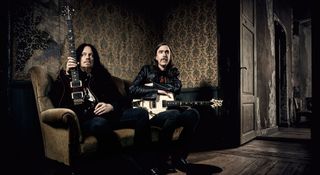 Opeth's Fredrik Åkesson [left] and Mikael Åkerfeldt sit on an old sofa with their PRS electric guitars. Åkesson looks off into the distance while Åkerfeldt stares into the camera.