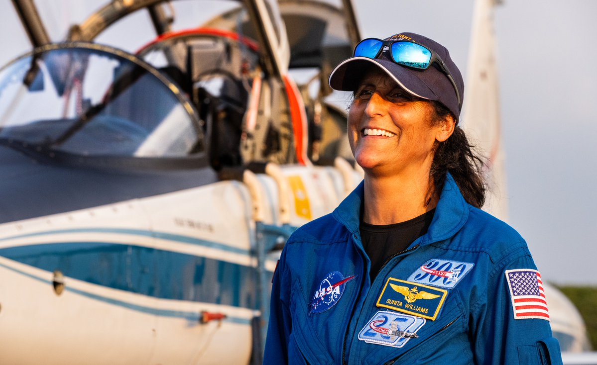 smiling woman astronaut in a flight suit and hat in front of a jet plane, with cockpit open