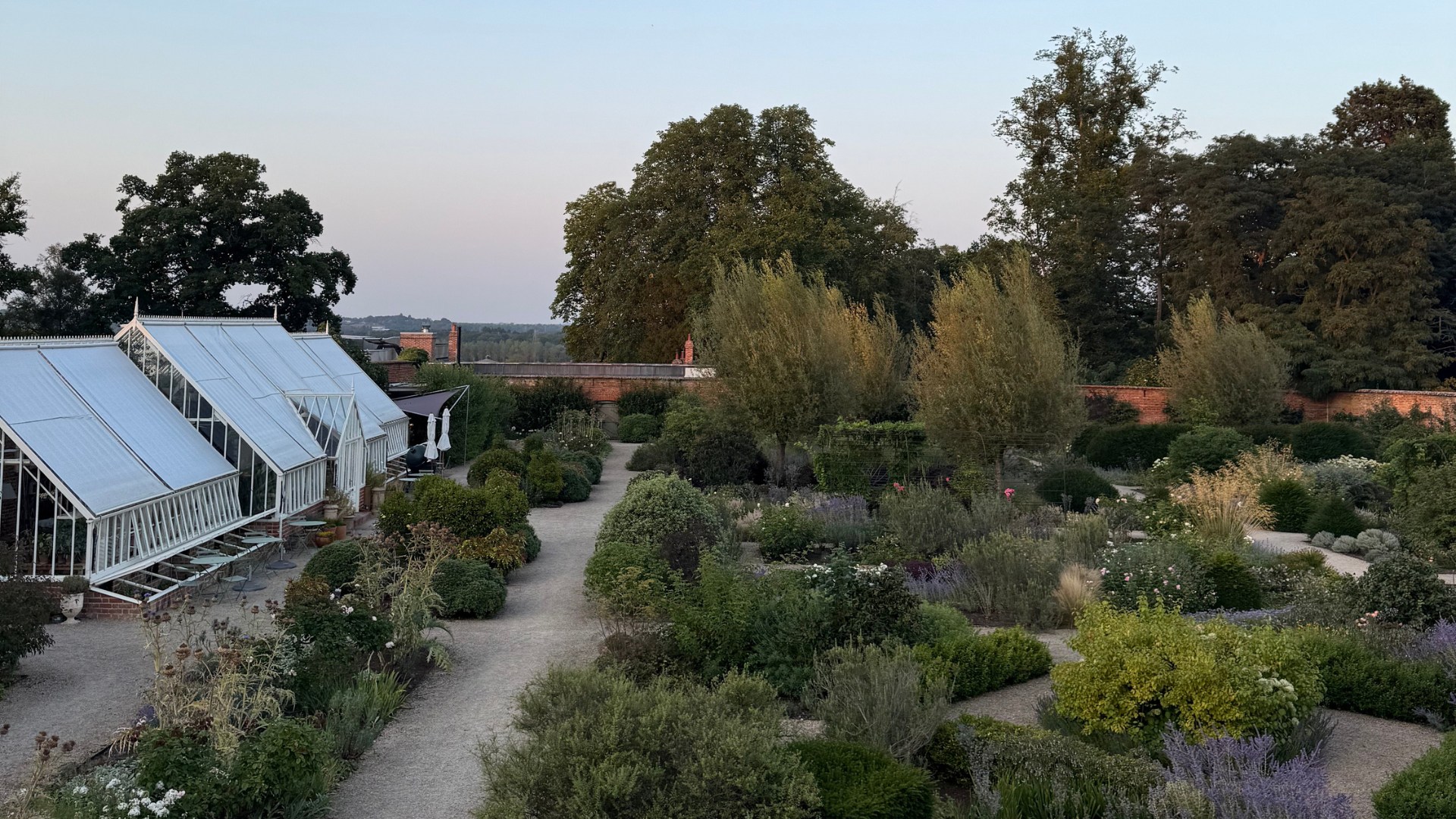 A walled garden at twilight, with gravel paths winding between neatly planted shrubs and borders. White glasshouses stand to the left, with tall trees rising behind the red-brick walls.
