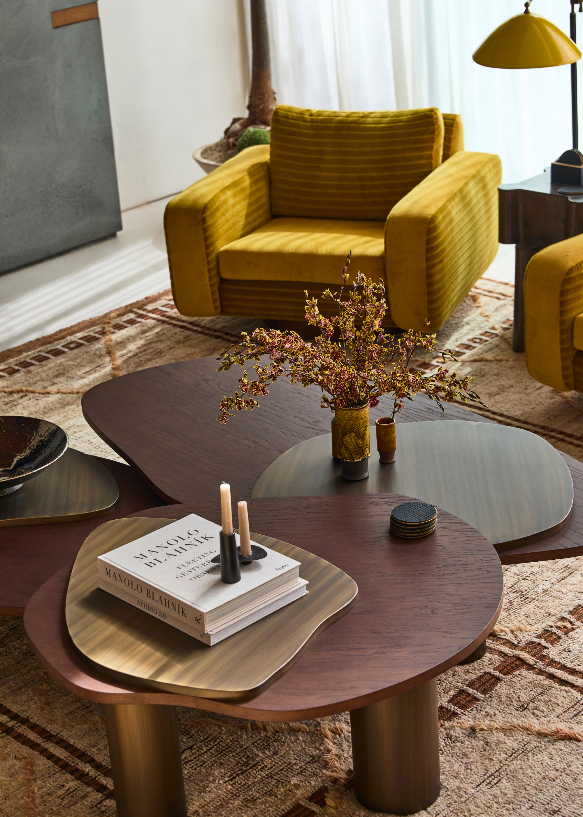 A trio of irregularly-shaped nesting coffee tables sit on top of a rug in a modern living room, styled with trays, books, candles and flowers with yellow armchairs in the background