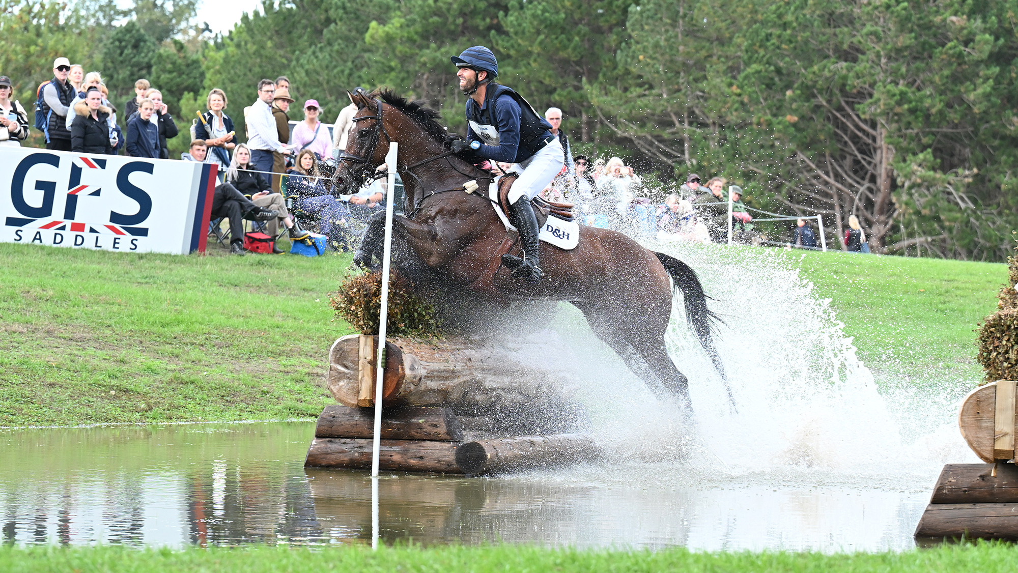 Clarke Johnstone, a rider in a blue top and hat, steers dark bay horse Sparky Lad over an angled fence in water at Blenheim Horse Trials 2026