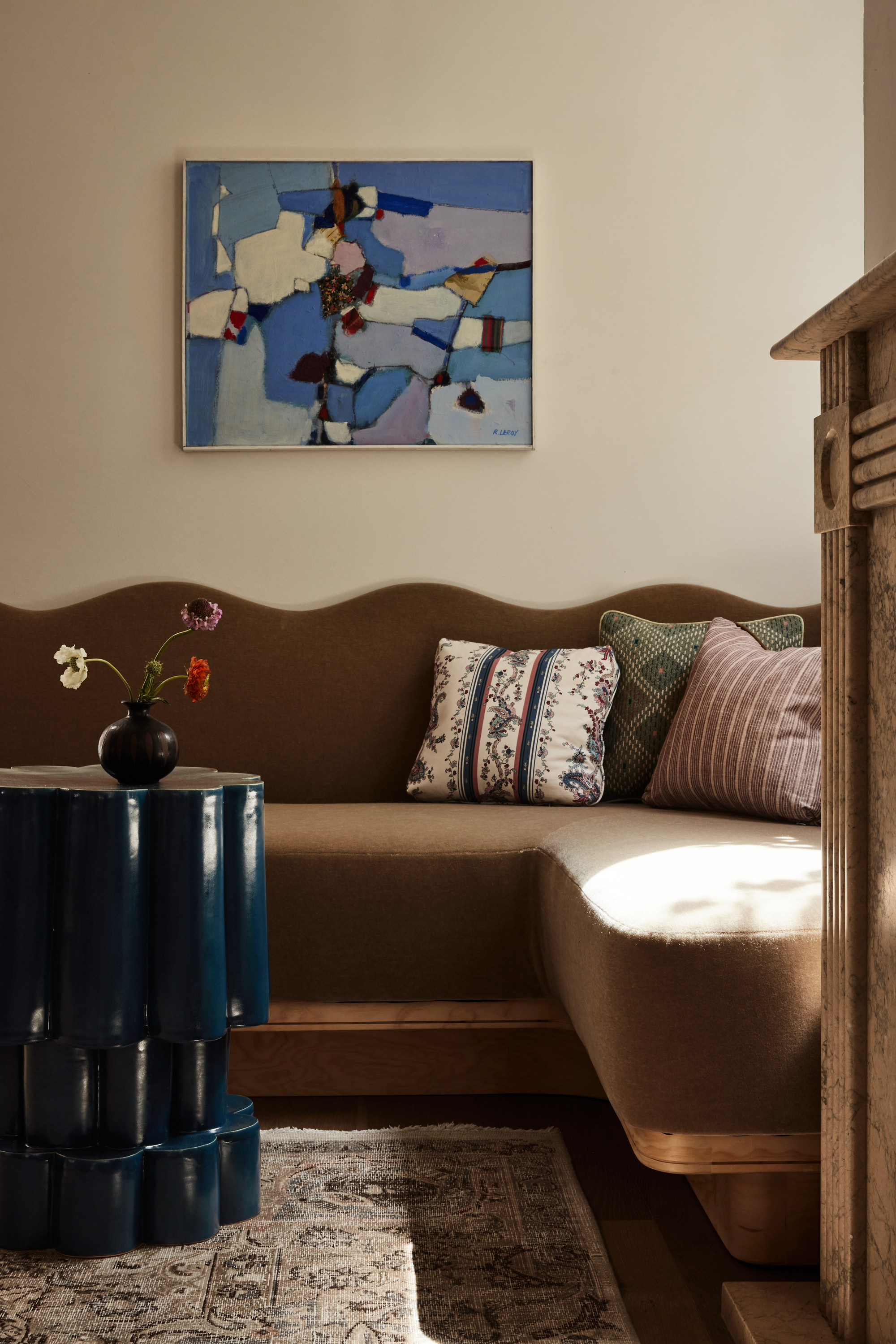 Image of a brown, velvet corner sofa with scalloped edges in a beige living room. There is a blue, abstract painting over the sofa, and a small, dark blue accent table in front of it.
