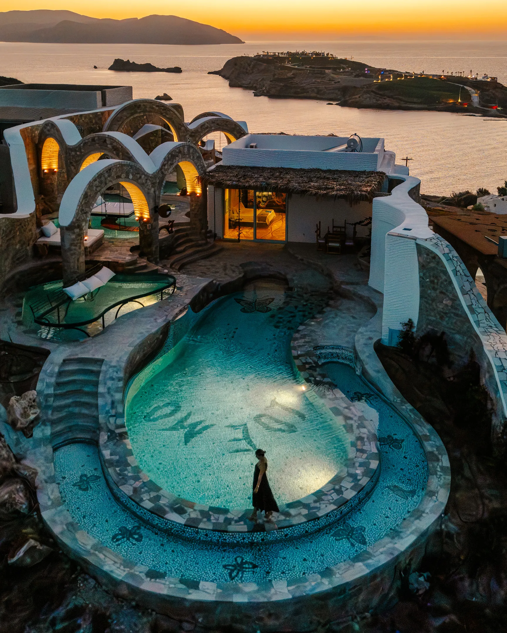 A mosaic pool with winding staircases and slides and butterly as well as floral-shaped details, caught just after golden hour from above as a woman dressed in a maxi dress walks its edge.