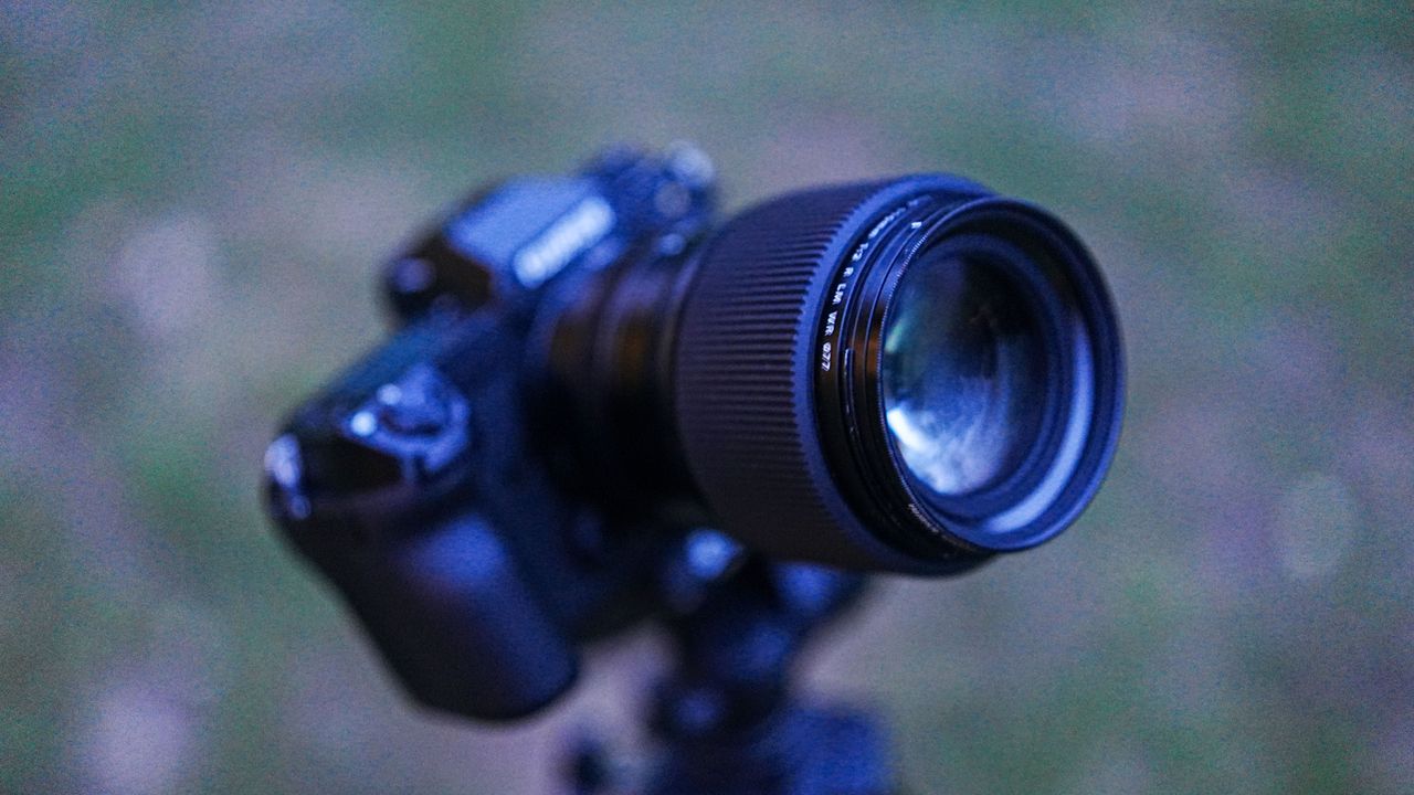 The Fujifilm GFX100S II with the Fujifilm GF 110mm f/2 R WR lens attached, on a tripod, aiming upward, with grass in the background.