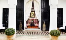 Exterior view of L’Hôtel, Marrakech, Morocco featuring black doors, white columns and patterned flooring. A red and white striped sofa, a painting and a table with flowers can be seen inside