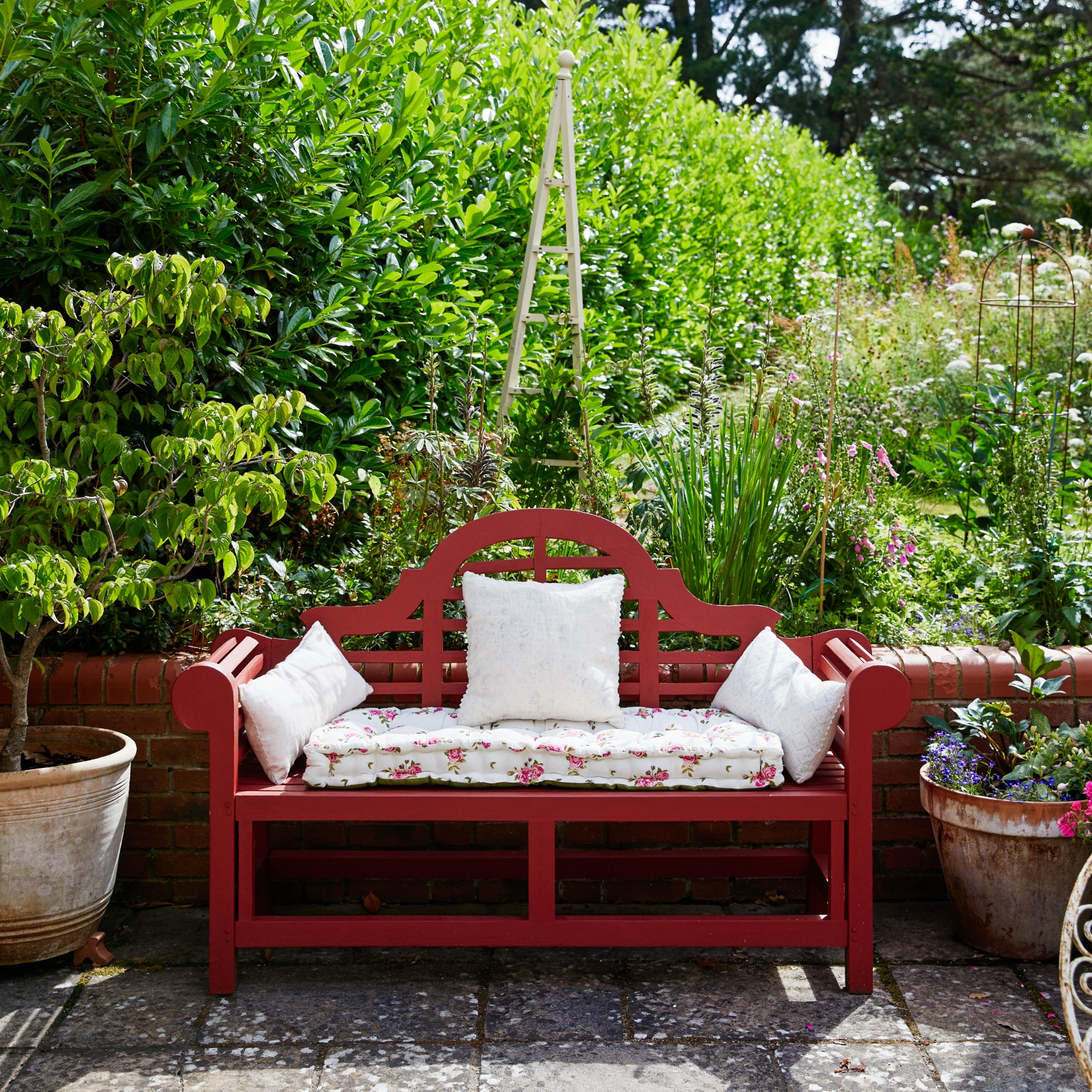 A red-painted curved wooden garden bench in a garden with flower beds and large terracorra plant pots