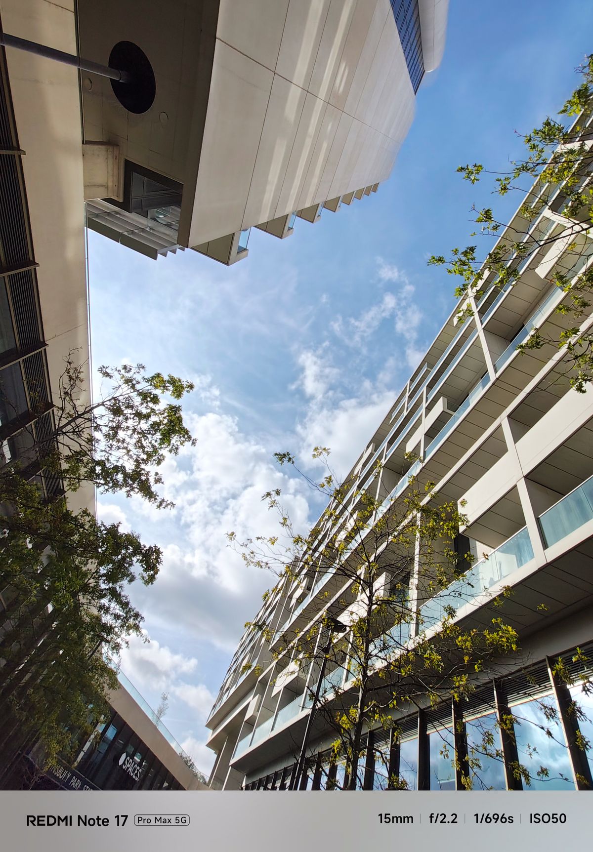 Upward wide-angle view between modern apartment buildings toward a blue and cloudy sky.