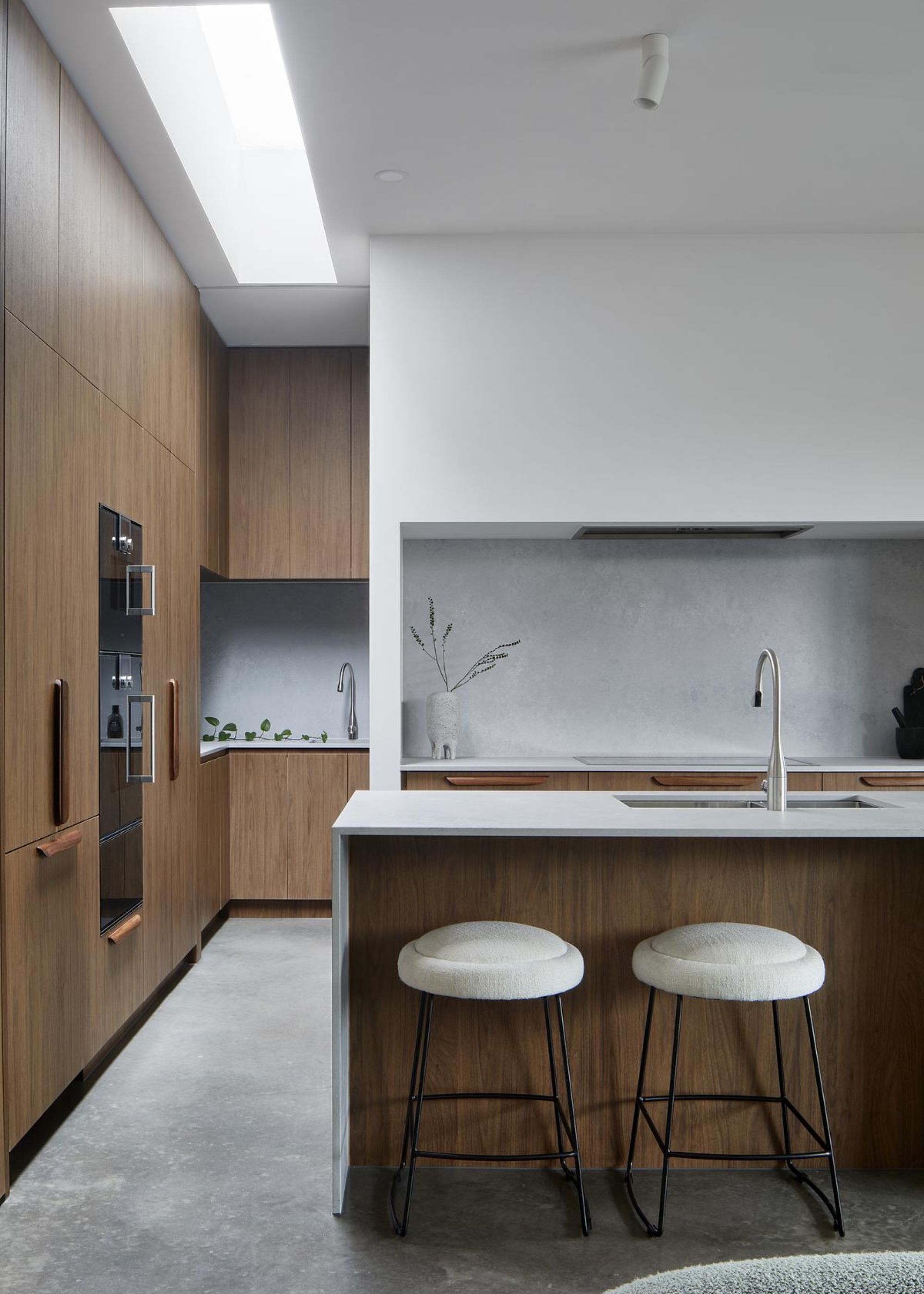A kitchen with an island in the middle as well as floor to ceiling cabinets. There is also a view of the corner of the kitchen with a pale white backsplash as well as the main area of the kitchen opposite the sink. Beside the kitchen island are two bar stools.