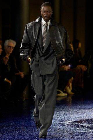 How to wear a suit is shown in a photo of a model on the runway of Saint Laurent's spring 2025 show wearing a black leather bomber jacker over a gray blazer that's styled with a white pinstripe button-down shirt, brown striped tie, gray trousers, gold cuff bracelets, and pointed pumps