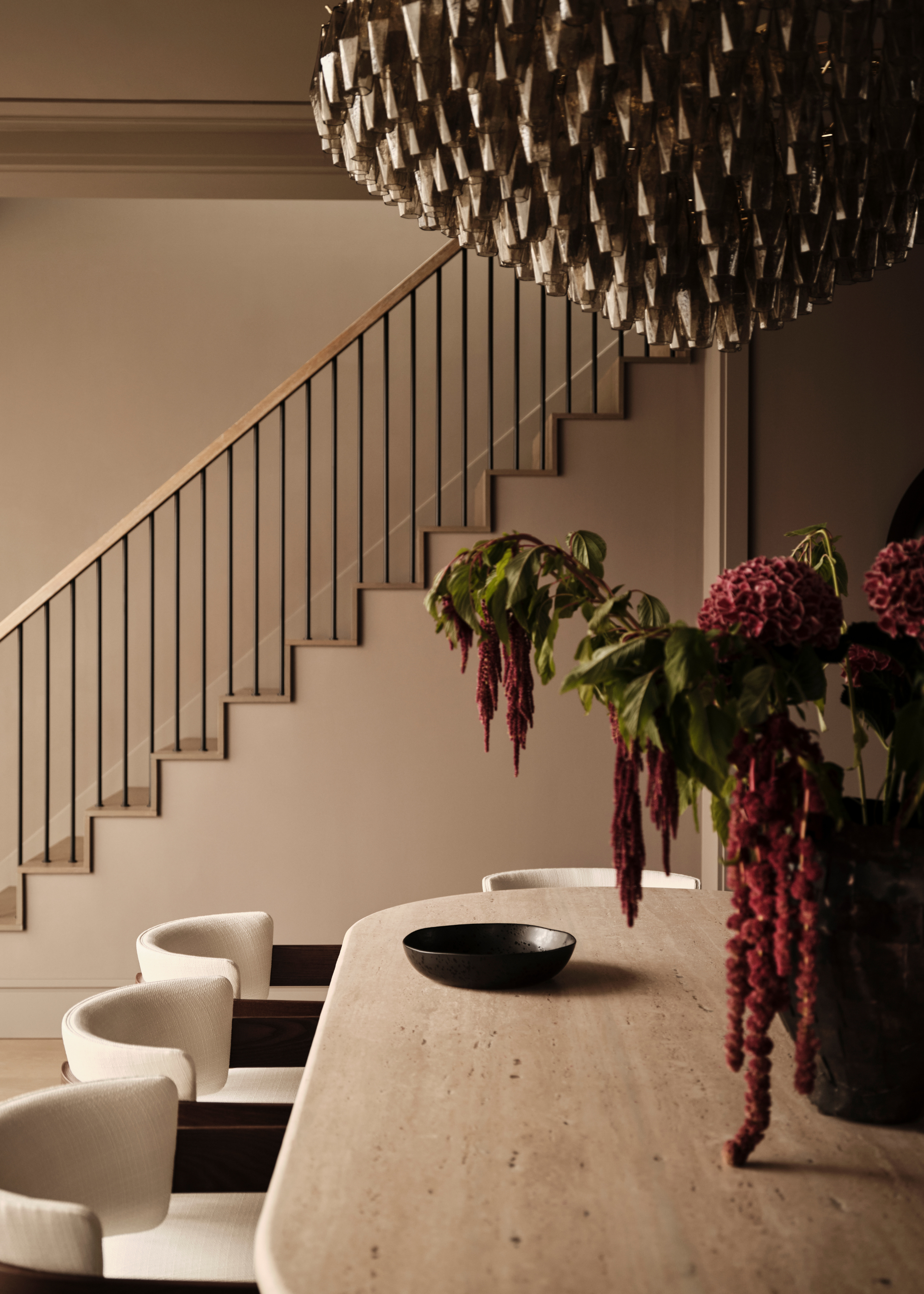 A natural hued dining table with curved edges and curved white-backed dining chairs with dark arm rests, with a large chanedelier hanging above the table with a floral display and a black bowl on top and a staircase in the background