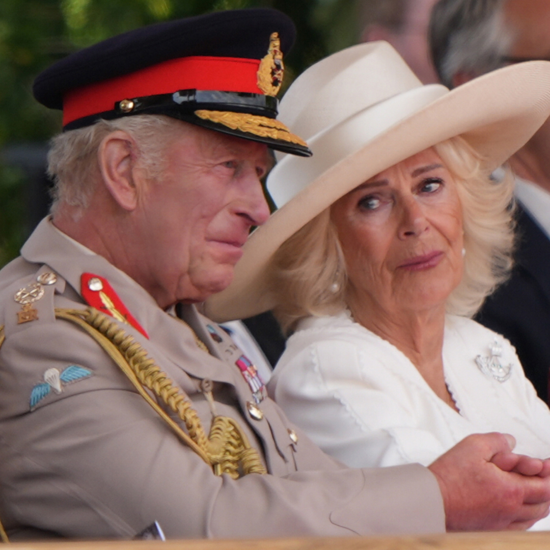King Charles wears a military uniform and Queen Camilla wears a white outfit and looks at him tearfully
