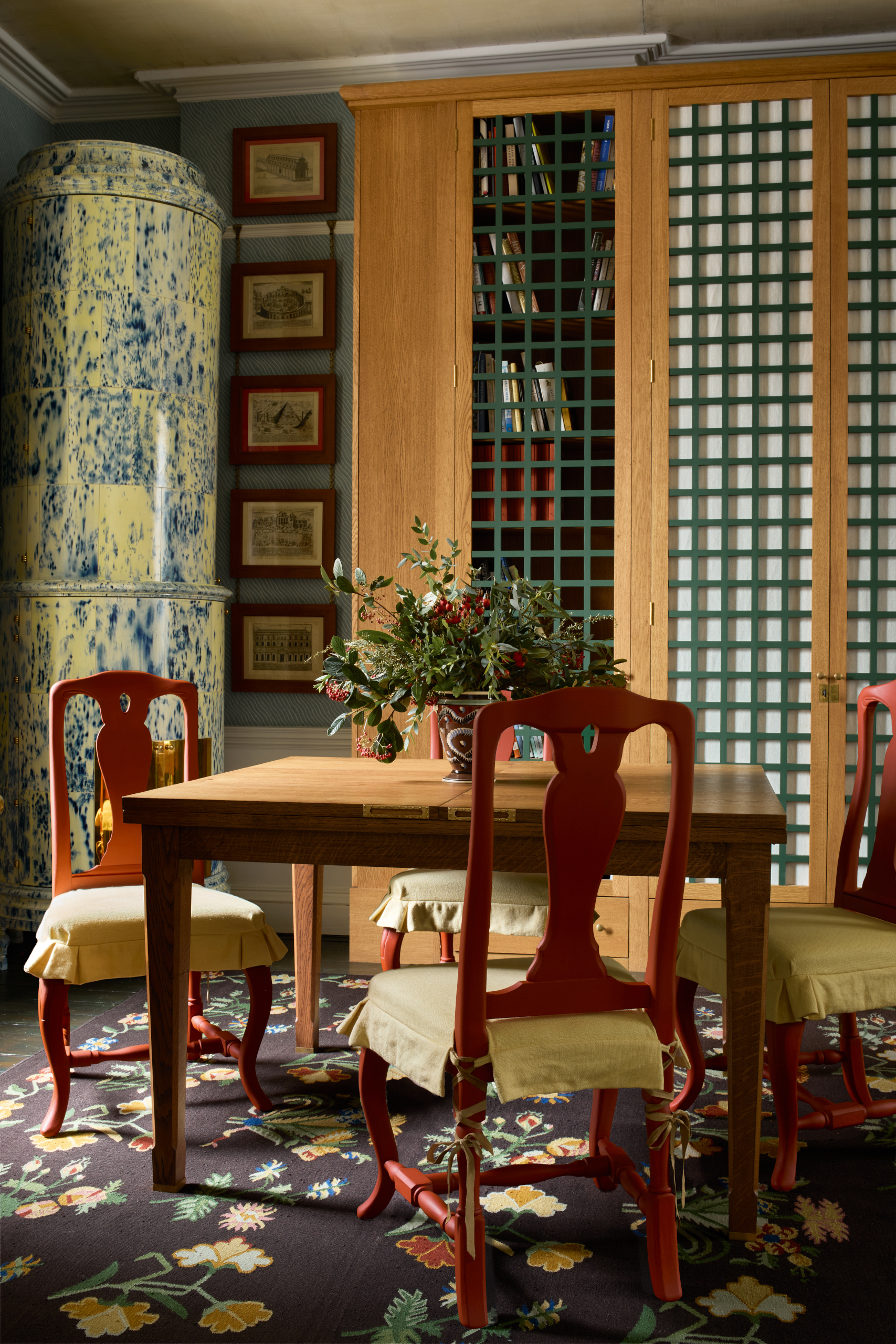 Green and blue kachelofen in a patterned room by Beata Heuman with red dining chairs and square wooden table