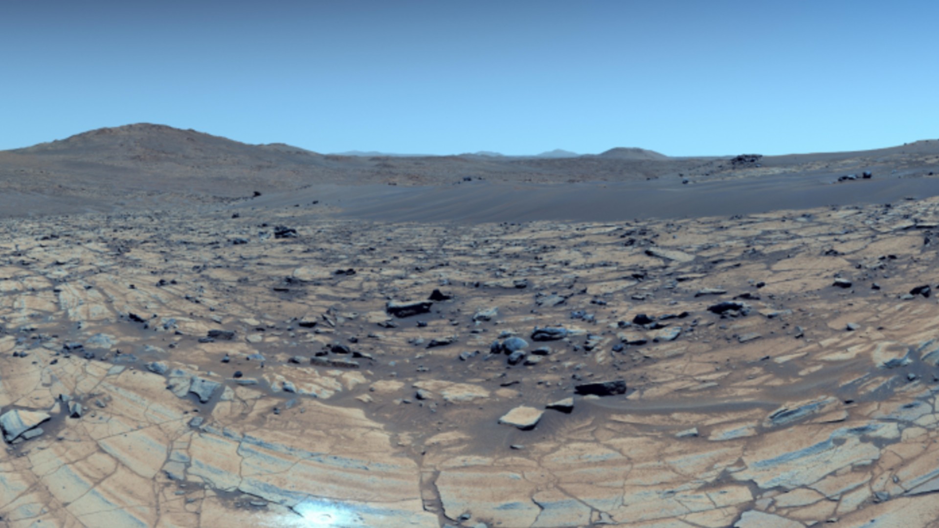 a reddish-orange landscape dotted with rocks of various sizes. mountains can be seen in background under a blue sky