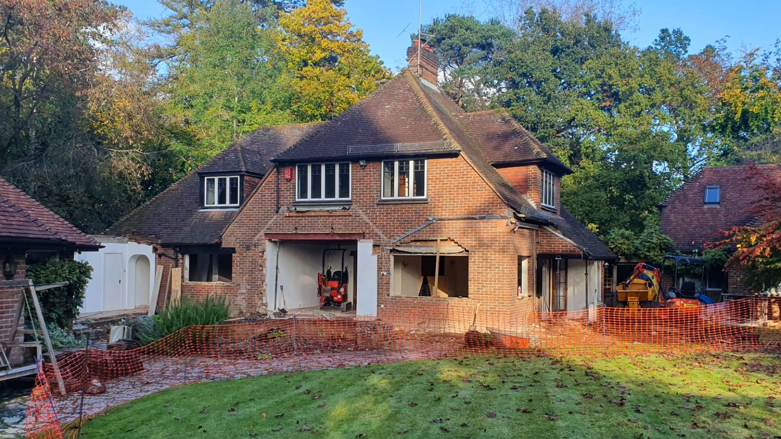 Red brick detached house with ground floor demolished, open to the elements, orange safety fencing across the garden, diggers visible inside.