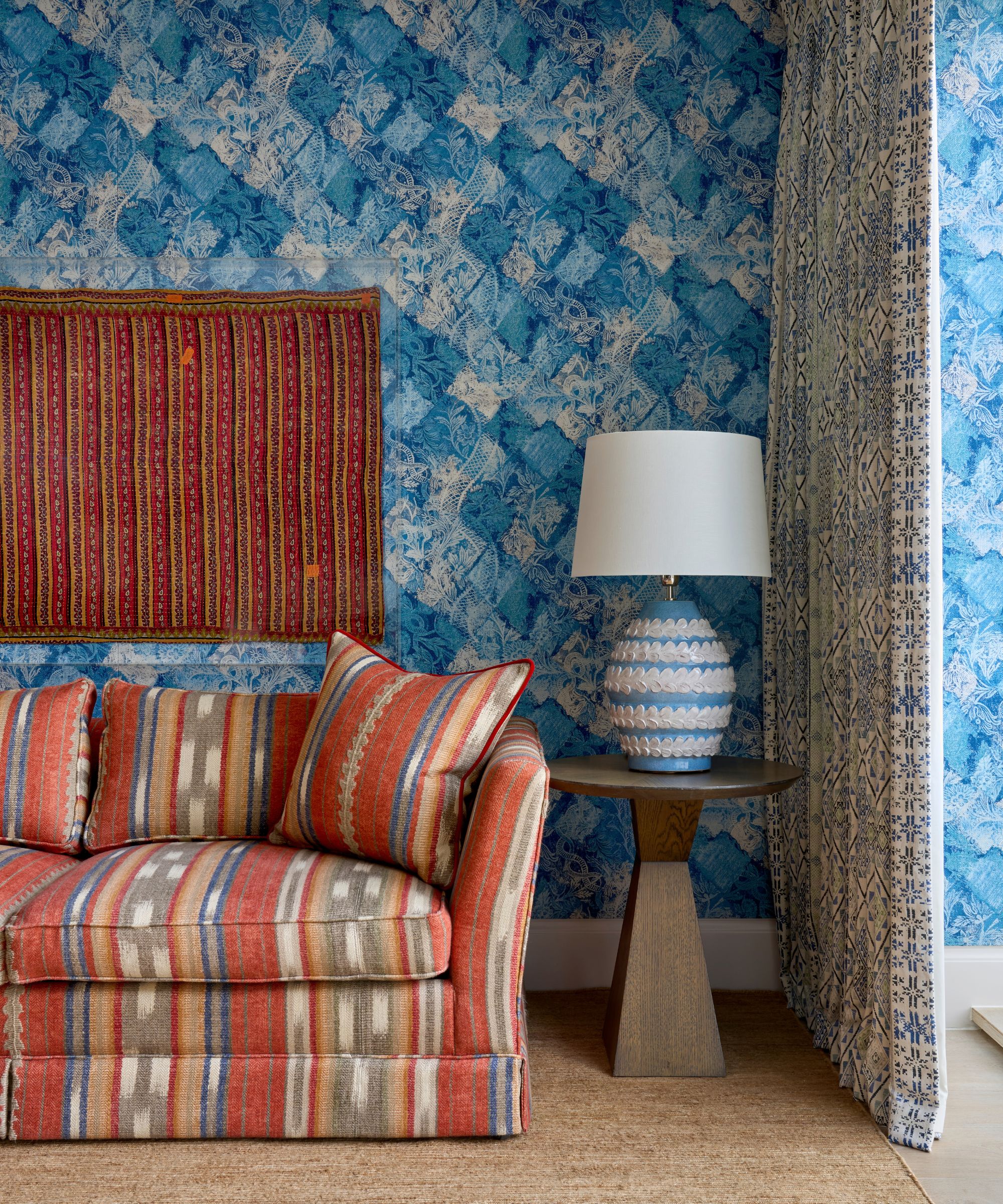 Living room with blue wallpaper, a red and yellow striped Ikat sofa, a side table with a large blue table lamp, a red fabric wall hanging, and a white and blue dividing curtain.