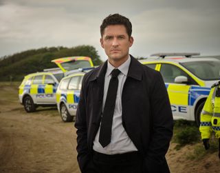 The Long Call - Ben Aldridge as DI Matthew Venn, in a shirt and tie with a long dark trenchcoat, standing on a Devon beach with police cars behind him