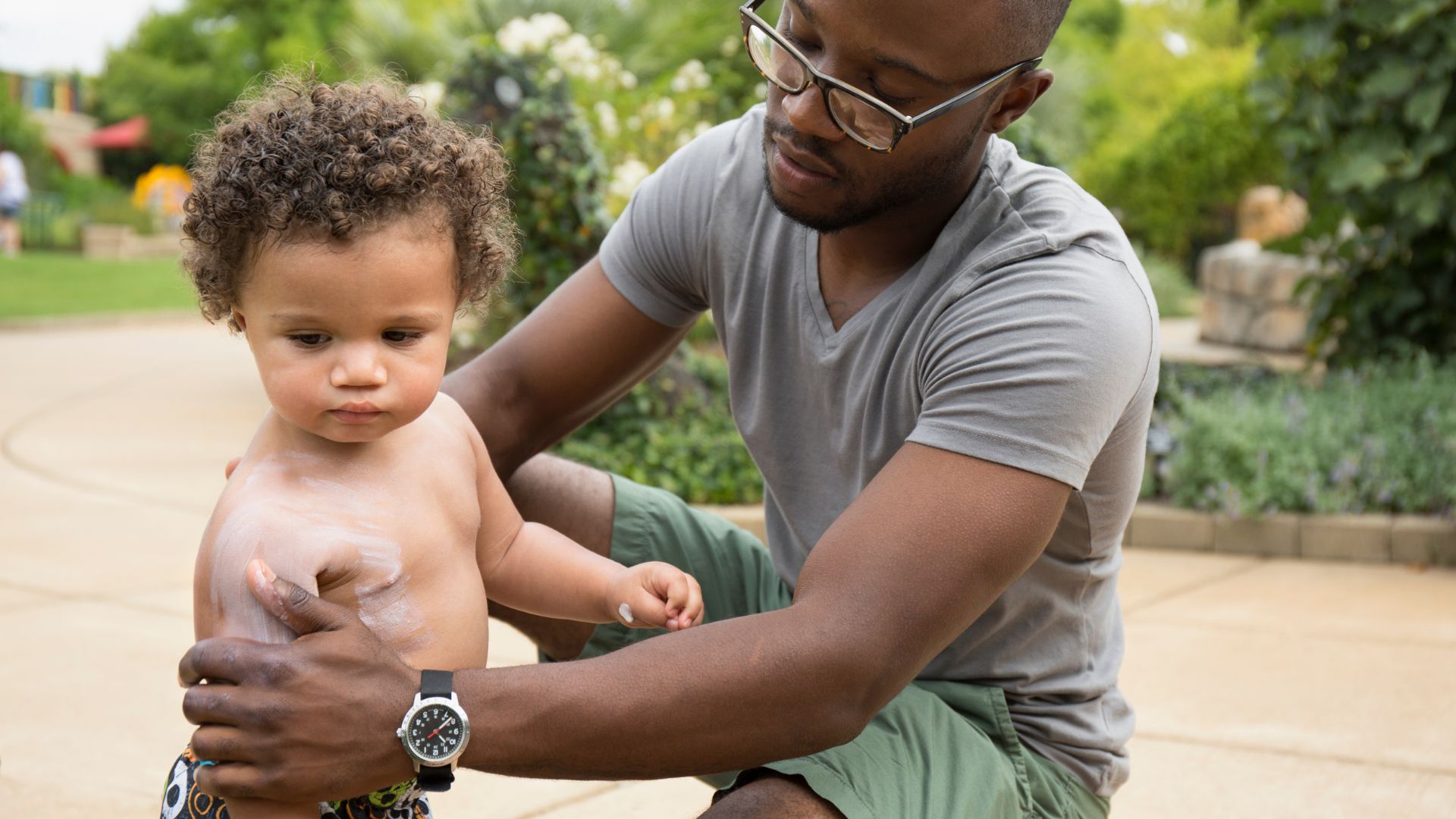 young father wearing glasses and a t shirt applies sunscreen to his toddler son's skin while both stand outside in a driveway