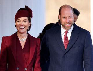 Kate Middleton wearing a maroon coat and hat and smiling standing next to Prince William wearing a blue coat and red tie