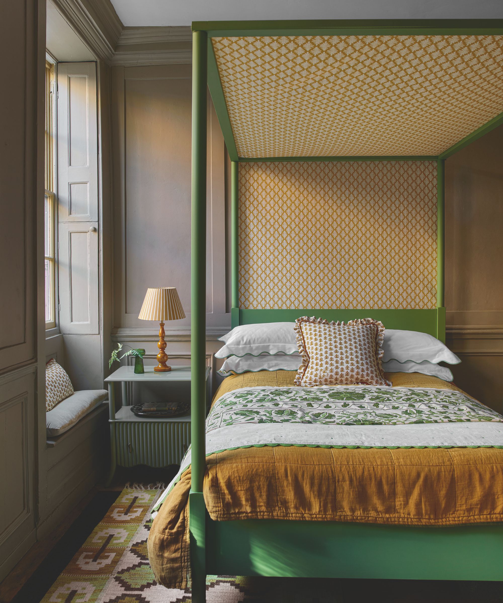 a bedroom with a green-painted four poster bed, yellow sheets, brown painted walls, a small side table, and a lamp on the table