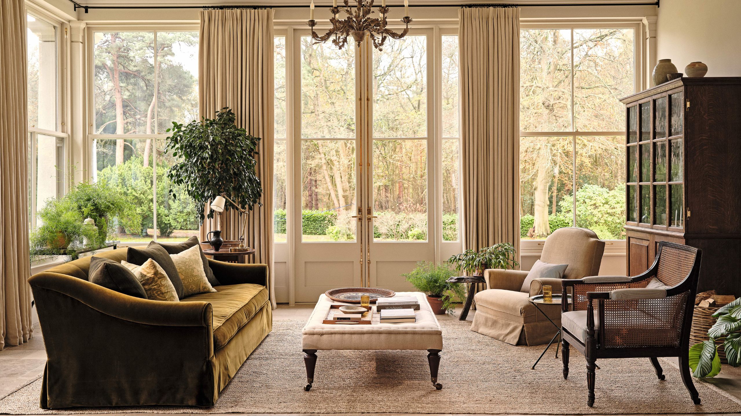 a large cozy sitting area in a grand orangery with french doors and windows styled with a green velvet sofa, ottoman in the middle, two mismatched arm chairs and a vintage bookcase