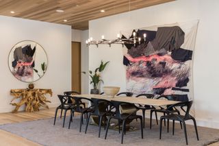 dining room with modern artwork pale wood ceiling cladding raw wooden sculptural console and black chairs round long pale wood table