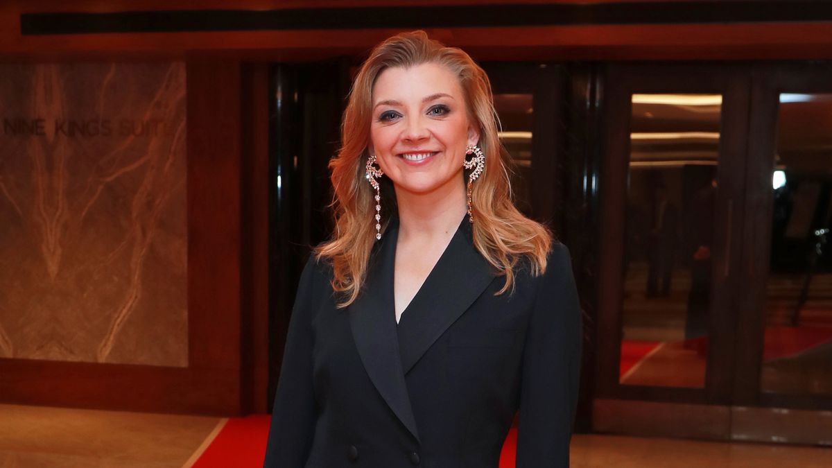 Natalie smiling as she stands in front of a hotel entrance wearing a smart black suit jacket and big earrings. 