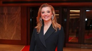 Natalie smiling as she stands in front of a hotel entrance wearing a smart black suit jacket and big earrings.