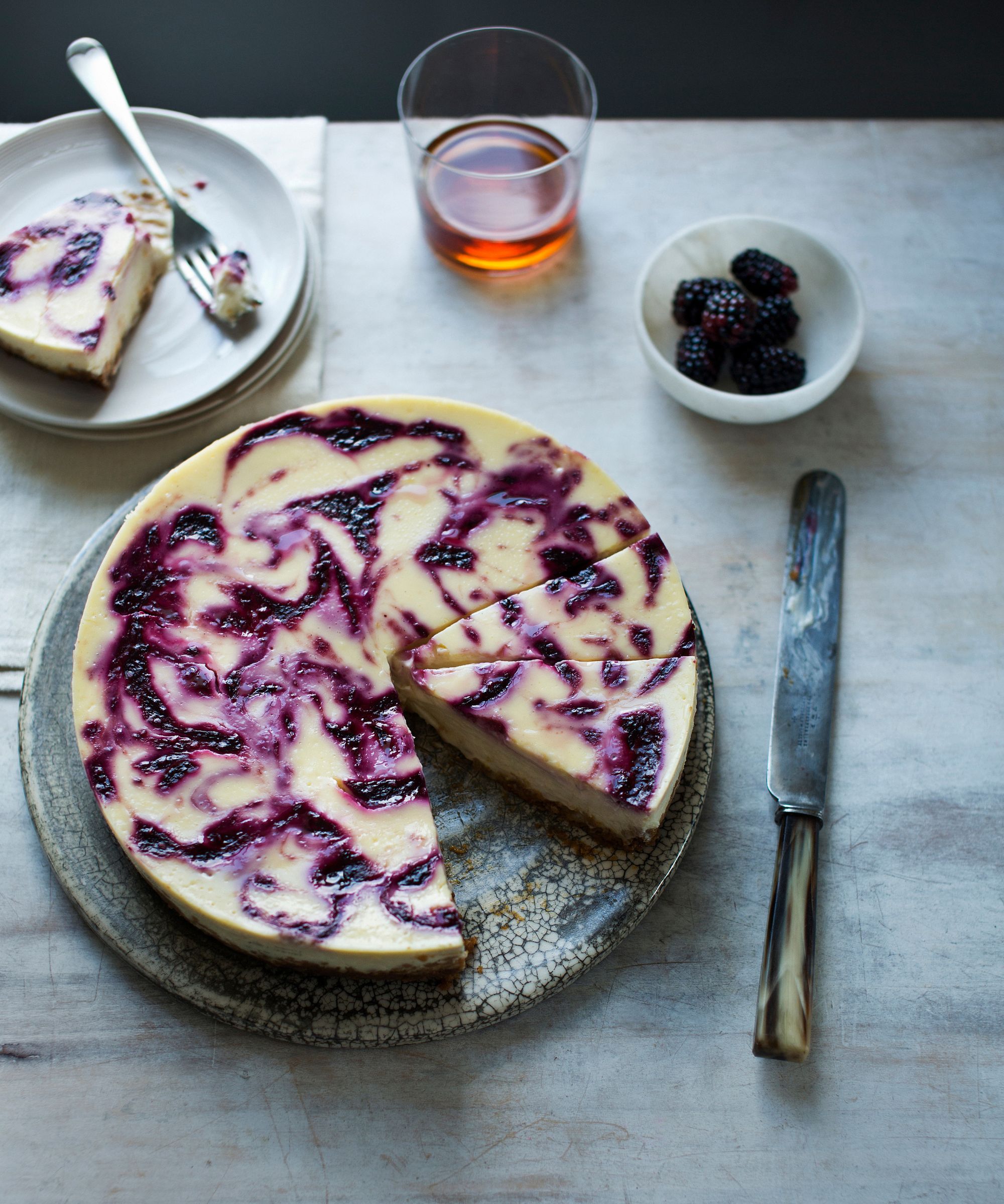 Blackberry ripple cheesecake on a plate with a slice on a separate place and a small bowl of blackberries and a knife to the side