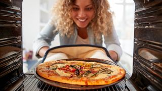 A woman putting a pizza in the oven with oven gloves and a tray