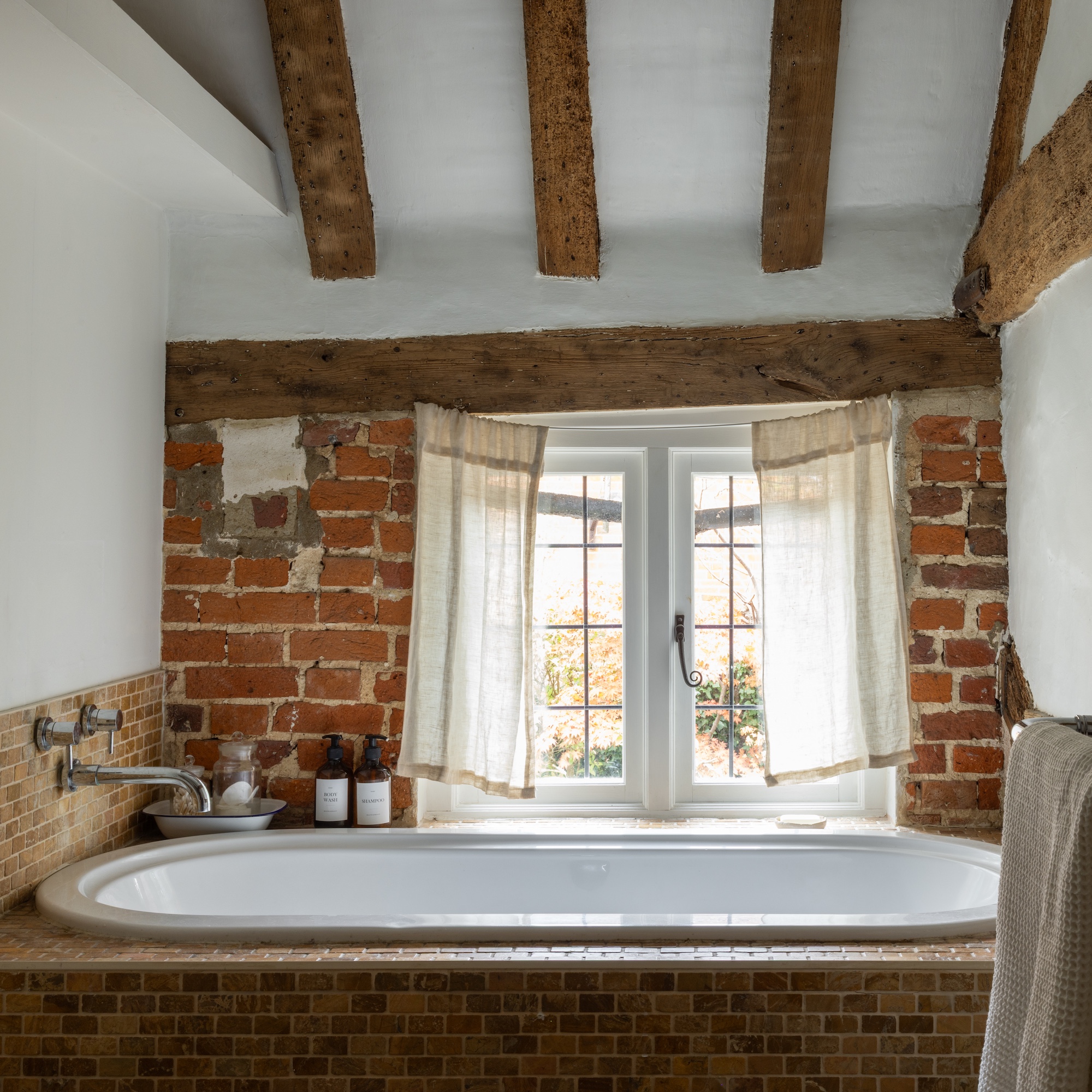 ensuite bathroom in old cottage with beamed ceiling and original brick walls, and bath with mosaic tile surround