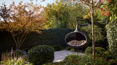 A corner of a modern garden with lots of hedges and flower beds, and a patio area with a black hanging egg chair with beige cushions inside. 