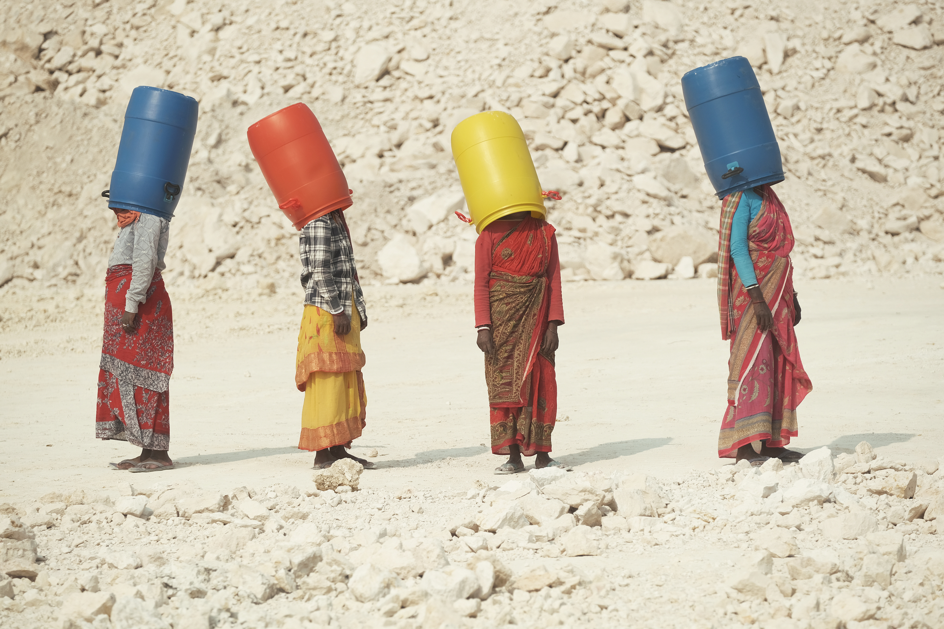 Four women in colourful saris stand in a line against a rocky, pale landscape, each balancing a large plastic barrel &amp;mdash; blue, orange, yellow, and blue &amp;mdash; on their head, obscuring their faces.